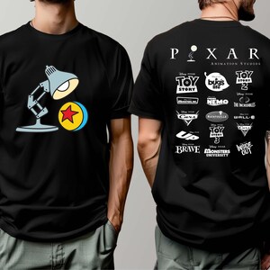May include: Black t-shirt featuring a cartoon illustration of the Pixar lamp and ball on the front. The back of the shirt displays the Pixar Animation Studios logo and logos from various Pixar films, including Toy Story, Finding Nemo, and Monsters University.
