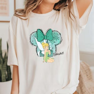 Pu&ograve; includere: T-shirt color crema con grafica di Trilli. Il design include un fiocco verde acqua a pois bianchi, una silhouette di fata e la scritta "Tinkerbell" in corsivo. La maglietta ha maniche corte.