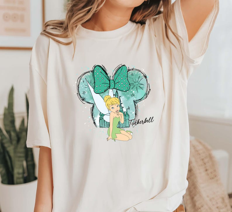 Pu&ograve; includere: T-shirt color crema con grafica di Trilli. Il design include un fiocco verde acqua a pois bianchi, una silhouette di fata e la scritta "Tinkerbell" in corsivo. La maglietta ha maniche corte.
