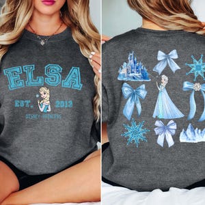 以下が含まれることがあります： ライトブルーの文字で「ELSA」と書かれたダークグレーのスウェットシャツ。エルサの画像と「Est. 2013 Disney Princess」の文字。背中にはエルサ、リボン、雪の結晶、青いお城が描かれています。