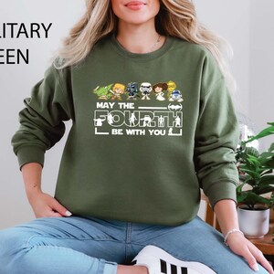 Puede incluir: Sudadera verde militar con un gráfico con personajes de Star Wars y el texto "MAY THE FOURTH BE WITH YOU". El diseño incluye versiones de dibujos animados de personajes icónicos. La sudadera es de cuello redondo.