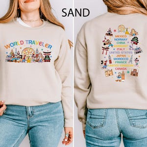 Puede incluir: Sudadera de color arena con el texto "WORLD TRAVELER" en letras arcoíris. La parte delantera presenta un gráfico colorido de varios monumentos y personajes de dibujos animados. La parte trasera muestra un diseño circular con nombres de países e ilustraciones.