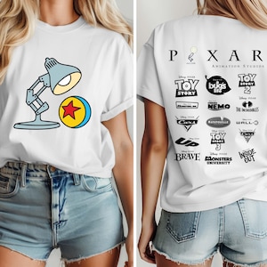 May include: White t-shirt featuring a cartoon image of a desk lamp and a yellow ball with a red star. The back of the shirt displays the Pixar Animation Studios logo and various movie titles in black.