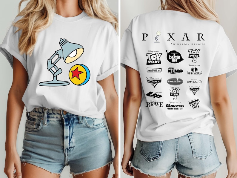 May include: White t-shirt featuring a cartoon image of a desk lamp and a yellow ball with a red star. The back of the shirt displays the Pixar Animation Studios logo and various movie titles in black.