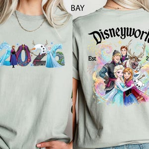 K&ouml;nnte beinhalten: Hellgr&uuml;nes T-Shirt mit farbenfroher Grafik von Frozen-Charakteren und dem Jahr 2026. Auf der R&uuml;ckseite des Shirts steht der Text "Disneyworld Est 2013" mit einer Gruppe von Charakteren.