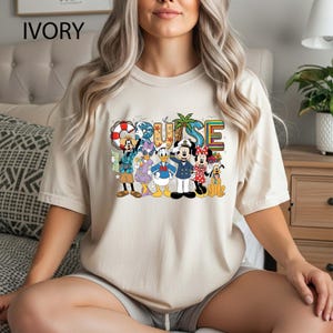 May include: Ivory-colored t-shirt featuring a colorful graphic of Disney characters with the word "CRUISE" above them. The characters include Mickey Mouse, Minnie Mouse, Donald Duck, and Goofy. The shirt is a crew neck and has short sleeves.