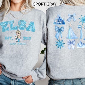 以下が含まれることがあります： ライトグレーのスウェットシャツ。前面には、水色の文字で「ELSA」、エルサのイラスト、そして「EST. 2013 DISNEY PRINCESS」の文字。背面には、リボン、雪の結晶、お城など、エルサをテーマにしたグラフィックが描かれています。