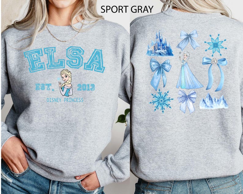 以下が含まれることがあります： ライトグレーのスウェットシャツ。前面には、水色の文字で「ELSA」、エルサのイラスト、そして「EST. 2013 DISNEY PRINCESS」の文字。背面には、リボン、雪の結晶、お城など、エルサをテーマにしたグラフィックが描かれています。