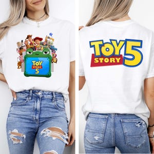 May include: A white t-shirt featuring a front graphic of Toy Story characters surrounding a green frog-shaped frame with the text Toy Story 5 inside. The back of the shirt displays the large blue and yellow text Toy Story 5 on a red background.