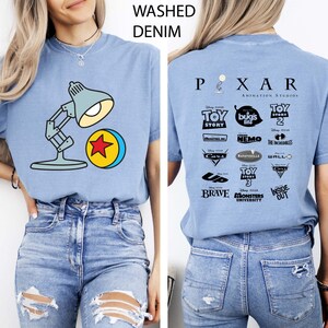 May include: Light blue t-shirt with a graphic of a desk lamp and a ball with a red star. The back of the shirt features the Pixar Animation Studios logo and various movie titles. The shirt is made of washed denim.