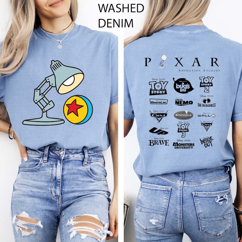 May include: Light blue t-shirt with a graphic of a desk lamp and a ball with a red star. The back of the shirt features the Pixar Animation Studios logo and various movie titles. The shirt is made of washed denim.