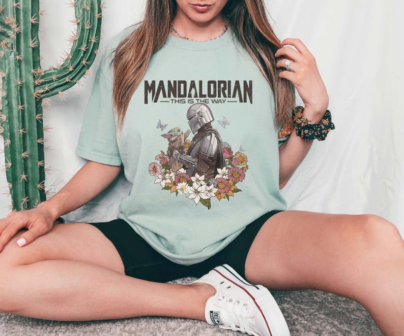 May include: A light green t-shirt featuring the text "MANDALORIAN - THIS IS THE WAY" in brown. The shirt has a graphic of the Mandalorian and Grogu surrounded by flowers and butterflies. The shirt is made of a soft material.