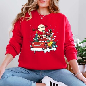 K&ouml;nnte beinhalten: Ein rotes Sweatshirt mit einem weihnachtlichen Motiv. Das Design zeigt ein rotes Auto mit Mickey und Minnie Mouse, Donald Duck und Goofy, vor Weihnachtsb&auml;umen und dem Schlitten des Weihnachtsmanns. Das Sweatshirt hat eine einfarbige rote Farbe.