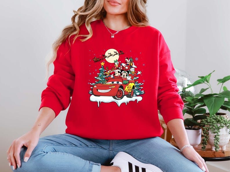 K&ouml;nnte beinhalten: Ein rotes Sweatshirt mit einem weihnachtlichen Motiv. Das Design zeigt ein rotes Auto mit Mickey und Minnie Mouse, Donald Duck und Goofy, vor Weihnachtsb&auml;umen und dem Schlitten des Weihnachtsmanns. Das Sweatshirt hat eine einfarbige rote Farbe.