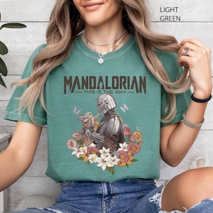 May include: Light green t-shirt featuring the text "MANDALORIAN" above an illustration of the Mandalorian and Grogu surrounded by flowers and butterflies. The phrase "THIS IS THE WAY" is below the word "MANDALORIAN".