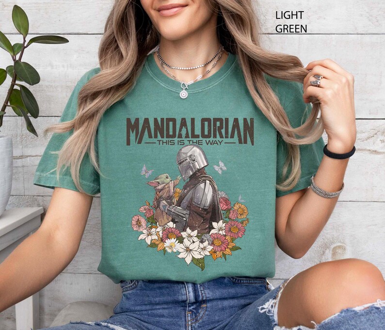 May include: Light green t-shirt featuring the text "MANDALORIAN" above an illustration of the Mandalorian and Grogu surrounded by flowers and butterflies. The phrase "THIS IS THE WAY" is below the word "MANDALORIAN".
