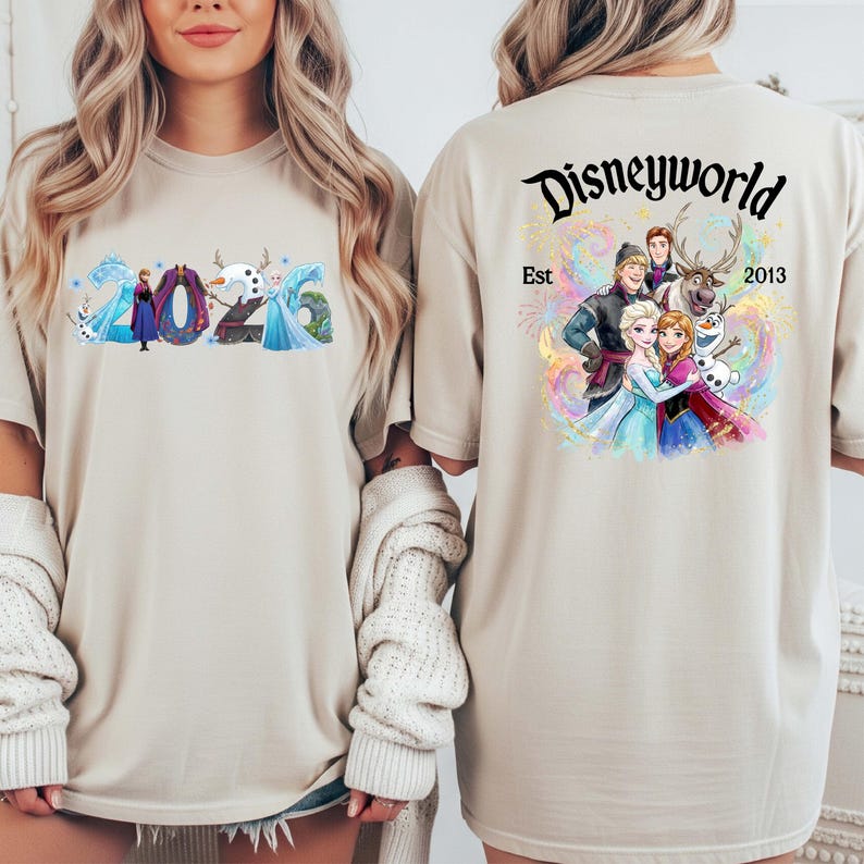 K&ouml;nnte beinhalten: Beiges T-Shirt mit Figuren aus dem Film Frozen. Auf der Vorderseite stehen die Zahlen "2026" mit den Figuren. Auf der R&uuml;ckseite des Shirts stehen die Worte "Disneyworld Est 2013" mit weiteren Figuren.