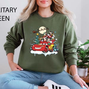 K&ouml;nnte beinhalten: Milit&auml;rgr&uuml;nes Sweatshirt mit einem festlichen Aufdruck von Disney-Figuren in einem roten Auto mit einem Weihnachtsbaum und einem Vollmond. Das Design umfasst Micky Maus, Minnie Maus, Donald Duck und Goofy. Der Text "MILITARY GREEN" steht oben.