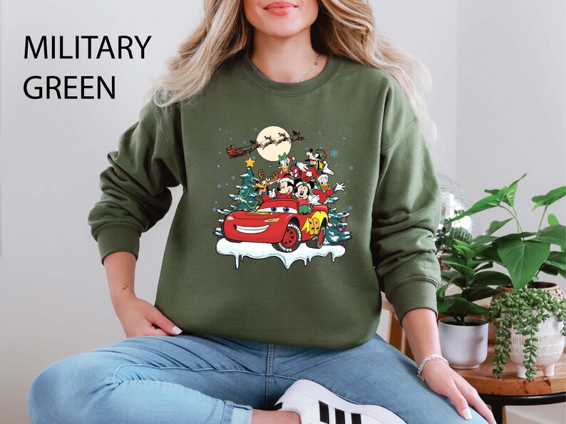 K&ouml;nnte beinhalten: Milit&auml;rgr&uuml;nes Sweatshirt mit einem festlichen Aufdruck von Disney-Figuren in einem roten Auto mit einem Weihnachtsbaum und einem Vollmond. Das Design umfasst Micky Maus, Minnie Maus, Donald Duck und Goofy. Der Text "MILITARY GREEN" steht oben.