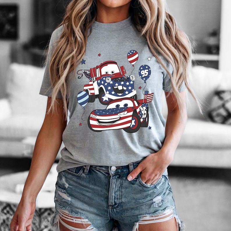 May include: A heather gray short-sleeve t-shirt featuring a graphic print of two cartoon cars decorated with red, white, and blue American flag patterns, stars, and balloons. One car is a tow truck and the other is a race car with the number 95.