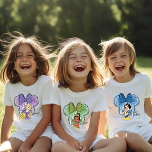 Pu&ograve; includere: Tre bambini indossano magliette bianche con disegni di principesse Disney. Le magliette presentano una silhouette di orecchio di Topolino con un'immagine di principessa e nome: Rapunzel, Tiana e Biancaneve. I bambini sorridono e sono seduti fuori.