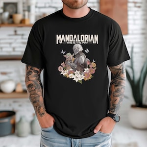 May include: Black t-shirt featuring a graphic of the Mandalorian and Grogu surrounded by flowers and butterflies. The text "MANDALORIAN - THIS IS THE WAY" is printed across the top in white.