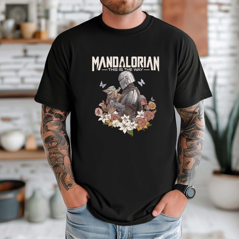May include: Black t-shirt featuring a graphic of the Mandalorian and Grogu surrounded by flowers and butterflies. The text "MANDALORIAN - THIS IS THE WAY" is printed across the top in white.