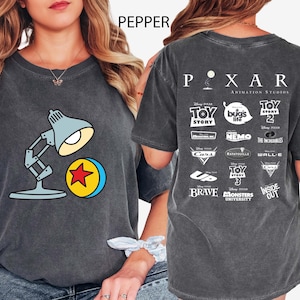 May include: Dark grey t-shirt featuring a cartoon lamp and ball design. The back of the shirt displays the Pixar Animation Studios logo and various movie titles. The word "PEPPER" is printed at the top.