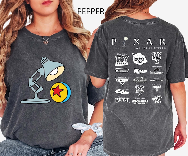 May include: Dark grey t-shirt featuring a cartoon lamp and ball design. The back of the shirt displays the Pixar Animation Studios logo and various movie titles. The word "PEPPER" is printed at the top.