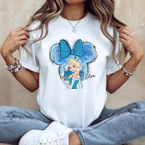 Pu&ograve; includere: T-shirt bianca con un'illustrazione a cartoni animati di Elsa di Frozen. Il design include un fiocco blu a pois bianchi, un castello e il nome "Elsa" in corsivo. Il design &egrave; all'interno di un contorno di orecchio di Topolino.