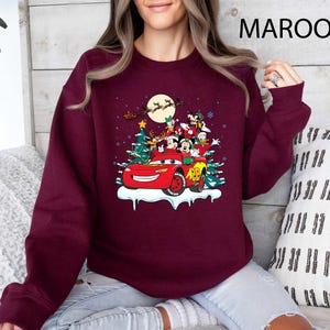 K&ouml;nnte beinhalten: Burgunderfarbenes Sweatshirt mit einem festlichen Disney-Motiv. Das Design zeigt Mickey Mouse, Donald Duck und andere Charaktere in einem roten Auto mit Weihnachtsbaum und Schnee. Der Text "MAROON" ist oben rechts aufgedruckt.