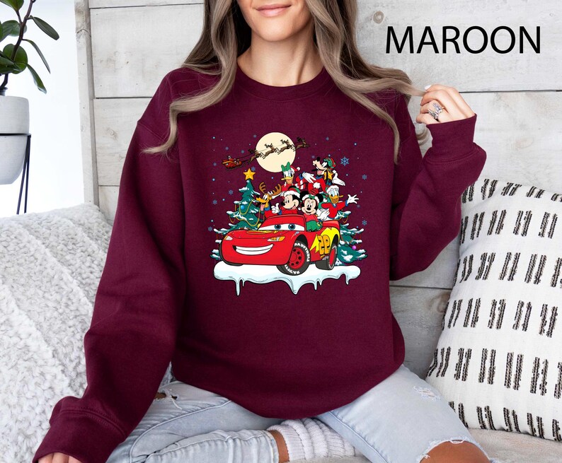 K&ouml;nnte beinhalten: Burgunderfarbenes Sweatshirt mit einem festlichen Disney-Motiv. Das Design zeigt Mickey Mouse, Donald Duck und andere Charaktere in einem roten Auto mit Weihnachtsbaum und Schnee. Der Text "MAROON" ist oben rechts aufgedruckt.