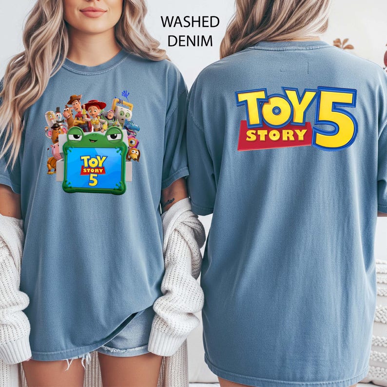 May include: A washed denim blue t-shirt featuring a front graphic of Toy Story characters surrounding a green frog-shaped frame with the text Toy Story 5. The back of the shirt displays the large yellow and red text Toy Story 5.