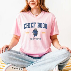May include: A light pink t-shirt featuring the text "CHIEF BOGO" in navy blue, with "EST. 2016" in gold. Below the text is an illustration of a bull in a police uniform, with the word "ZOOTOPIA" below. The shirt is made of a soft material.