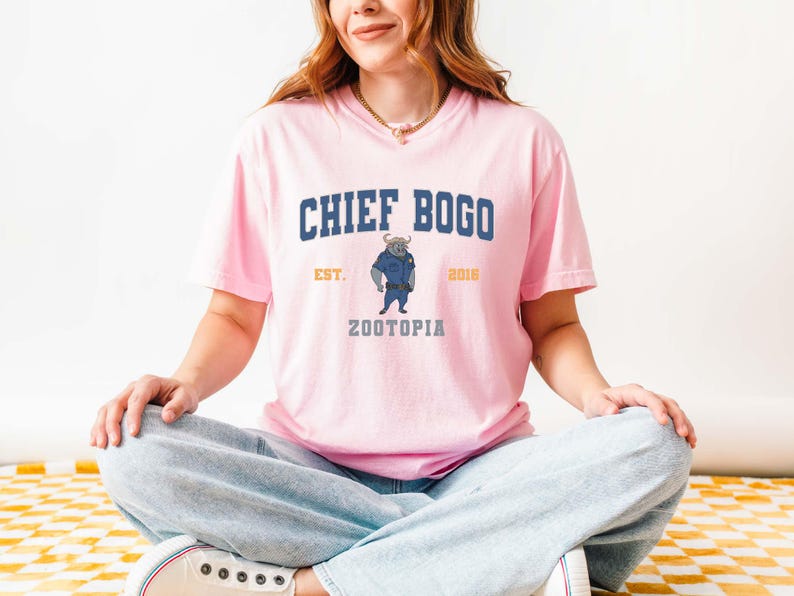 May include: A light pink t-shirt featuring the text "CHIEF BOGO" in navy blue, with "EST. 2016" in gold. Below the text is an illustration of a bull in a police uniform, with the word "ZOOTOPIA" below. The shirt is made of a soft material.
