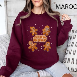 May include: A maroon sweatshirt with gingerbread cookie designs of Star Wars characters. The Star Wars logo is also displayed in a gingerbread cookie style. The sweatshirt is a solid maroon colour.