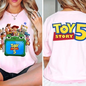 May include: A light pink t-shirt featuring a graphic of Toy Story characters emerging from a green frog-shaped box on the front, with the text Toy Story 5. The back of the shirt displays the large blue and yellow text Toy Story 5.