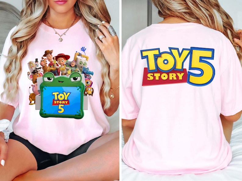 May include: A light pink t-shirt featuring a graphic of Toy Story characters emerging from a green frog-shaped box on the front, with the text Toy Story 5. The back of the shirt displays the large blue and yellow text Toy Story 5.