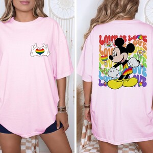 May include: Light pink t-shirt featuring a Mickey Mouse design with a rainbow heart made by hands on the front. The back of the shirt has a colorful Mickey Mouse graphic with the words "LOVE IS LOVE".