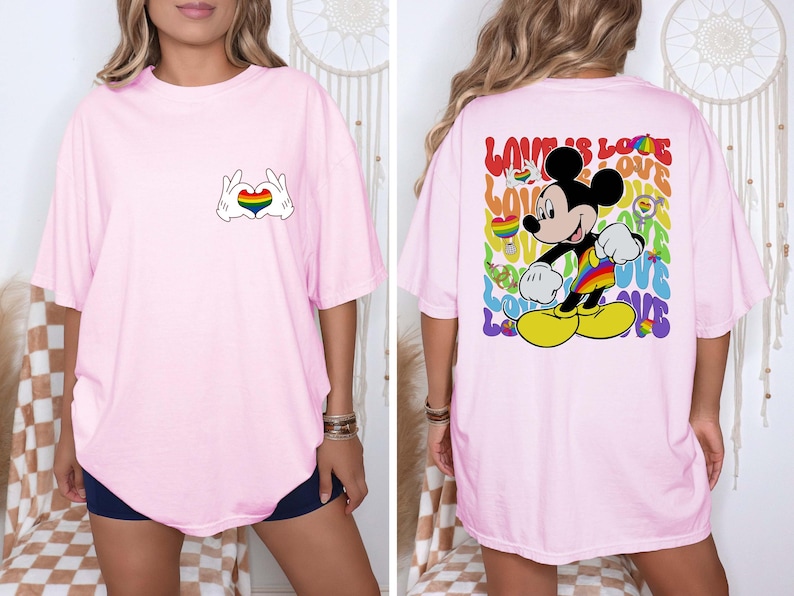 May include: Light pink t-shirt featuring a Mickey Mouse design with a rainbow heart made by hands on the front. The back of the shirt has a colorful Mickey Mouse graphic with the words "LOVE IS LOVE".