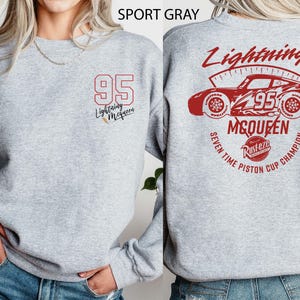 Może przedstawiać: Szara bluza sportowa z czerwoną grafiką Zygzaka McQueena i numerem 95. Z przodu widnieje numer 95 i napis "Lightning McQueen". Z tyłu znajduje się czerwona grafika samochodu, "Lightning McQueen" i napis "Seven Time Piston Cup Champion".