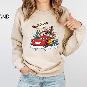 K&ouml;nnte beinhalten: Sandfarbenes Sweatshirt mit einem festlichen Aufdruck von Disney-Figuren in einem roten Auto mit Weihnachtsb&auml;umen und dem Schlitten des Weihnachtsmanns. Das Design umfasst Micky Maus, Donald Duck und Goofy. Das Wort "SAND" ist oben links aufgedruckt.