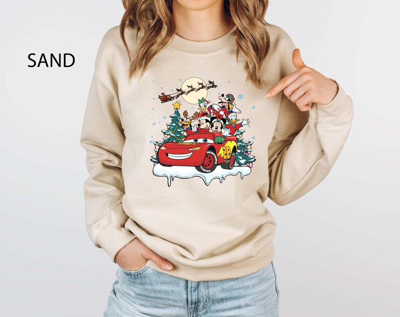 K&ouml;nnte beinhalten: Sandfarbenes Sweatshirt mit einem festlichen Aufdruck von Disney-Figuren in einem roten Auto mit Weihnachtsb&auml;umen und dem Schlitten des Weihnachtsmanns. Das Design umfasst Micky Maus, Donald Duck und Goofy. Das Wort "SAND" ist oben links aufgedruckt.