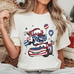 May include: A cream-colored t-shirt featuring a graphic print of two cartoon vehicles decorated with red, white, and blue American flag patterns. The design includes balloons, stars, and the number 95, with one vehicle resembling a tow truck.