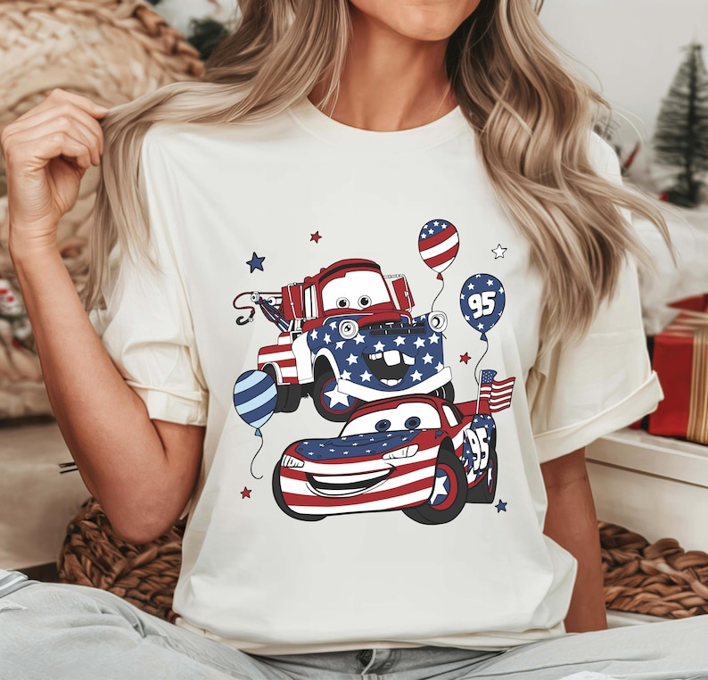 May include: A cream-colored t-shirt featuring a graphic print of two cartoon vehicles decorated with red, white, and blue American flag patterns. The design includes balloons, stars, and the number 95, with one vehicle resembling a tow truck.