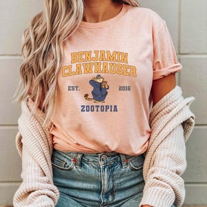 May include: A light pink t-shirt featuring the text "BENJAMIN CLAWHAUSER" in gold lettering above an illustration of a cartoon character in a police uniform. The text "EST. 2016 ZOOTOPIA" is also printed on the shirt.