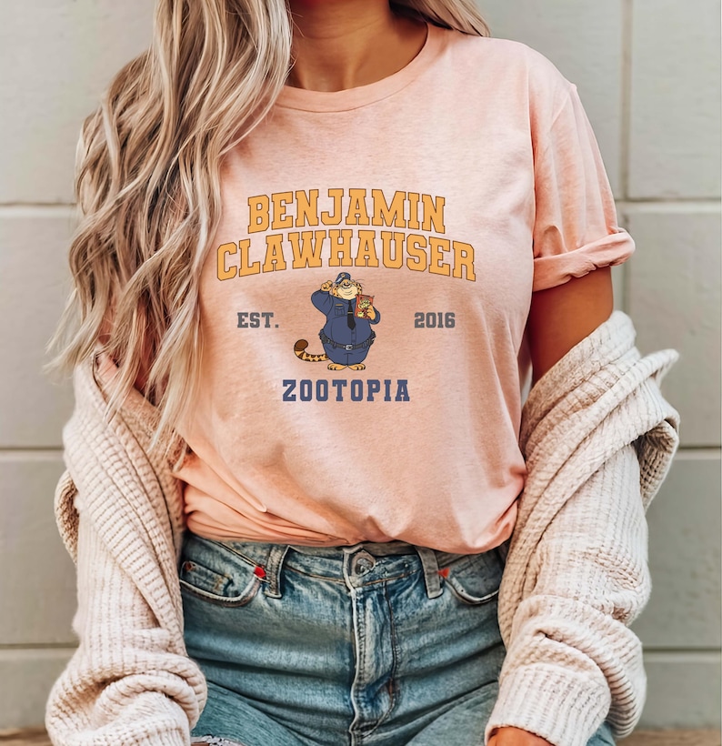 May include: A light pink t-shirt featuring the text "BENJAMIN CLAWHAUSER" in gold lettering above an illustration of a cartoon character in a police uniform. The text "EST. 2016 ZOOTOPIA" is also printed on the shirt.