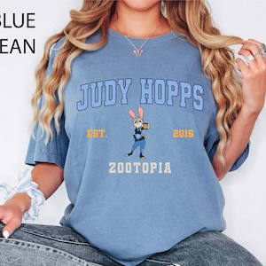 May include: A light blue t-shirt with the text "JUDY HOPPS" in a collegiate font, a cartoon image of Judy Hopps, and the text "ZOOTOPIA". The shirt also has the text "EST. 2016".