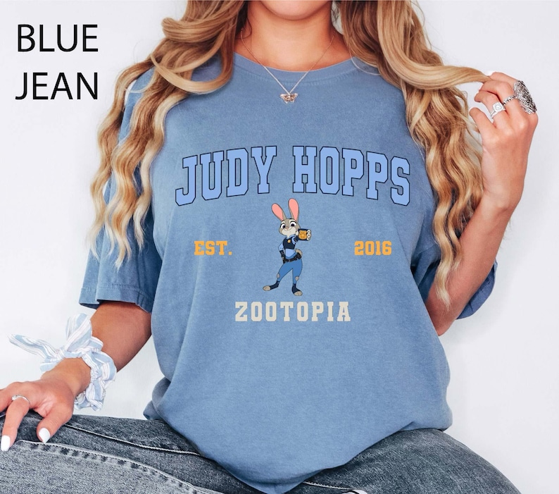 May include: A light blue t-shirt with the text "JUDY HOPPS" in a collegiate font, a cartoon image of Judy Hopps, and the text "ZOOTOPIA". The shirt also has the text "EST. 2016".