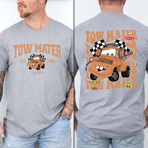Puede incluir: Una camiseta gris jaspeado con el personaje Tow Mater de la pel&iacute;cula Cars. La parte delantera de la camiseta tiene el texto "Tow Mater" y "Cars" con una imagen de Mater. La parte trasera de la camiseta tiene una imagen m&aacute;s grande de Mater con banderas a cuadros.